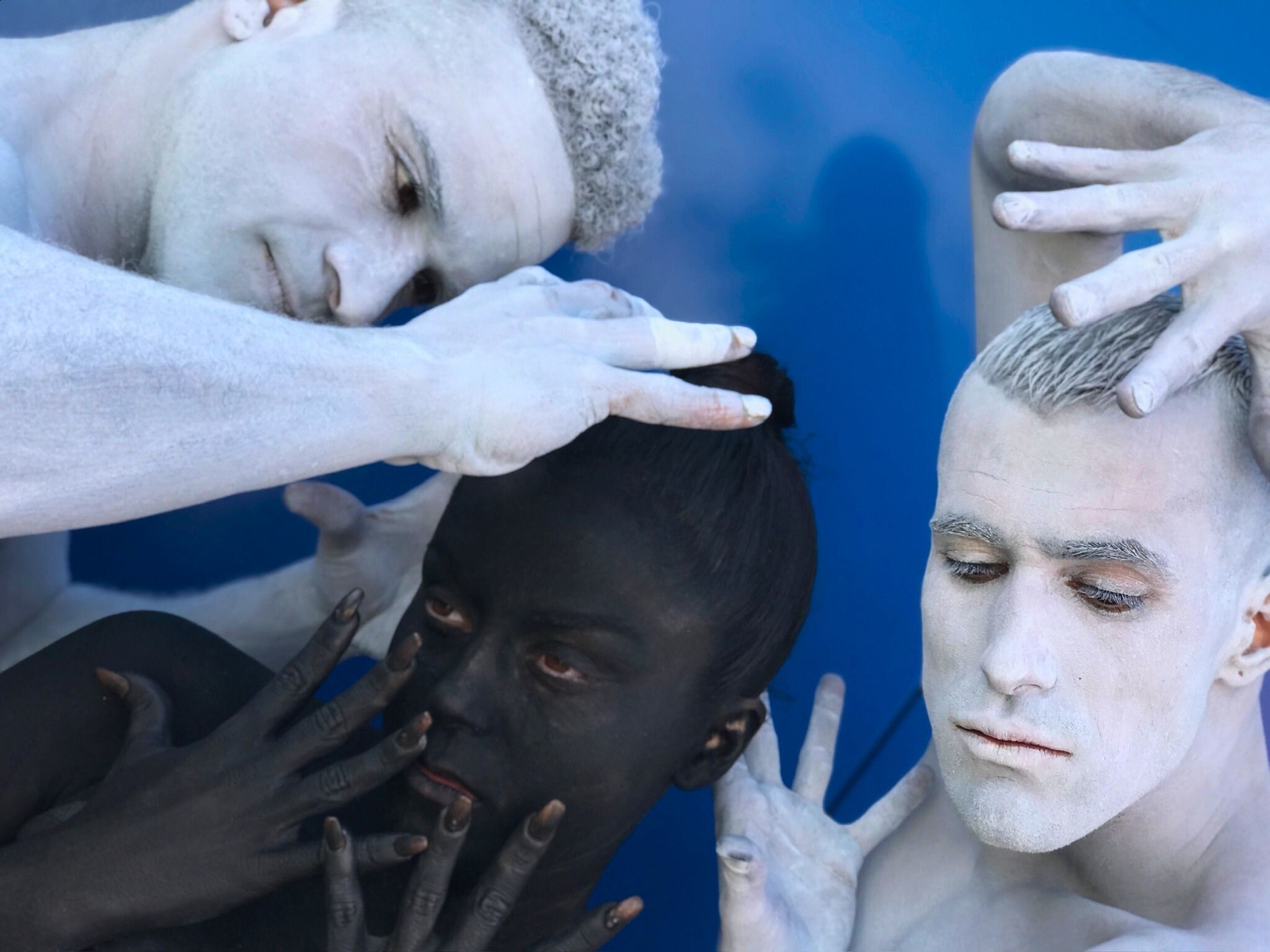 Víctor Óscar Juaranz, responsable de OjoArtísticoEvents, aparece caracterizado con el cuerpo y rostro pintados de blanco, a la derecha de la imagen, en una escena performativa junto a otros dos intérpretes sobre un fondo azul.