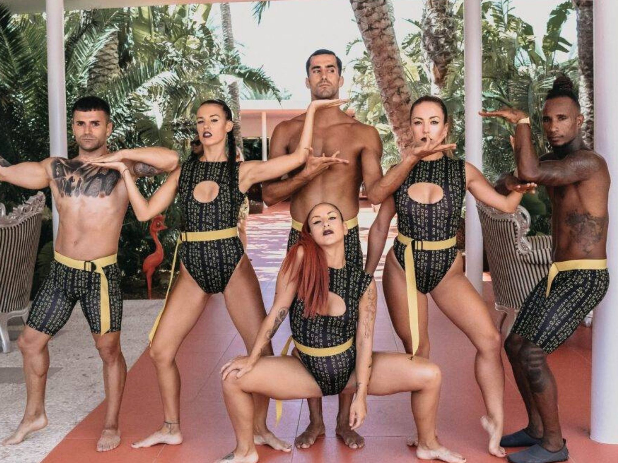 Grupo de seis artistas escénicos, incluyendo a Víctor Óscar Juaranz en el centro, posando en formación coreográfica bajo una pérgola tropical, con trajes coordinados de diseño moderno. Imagen de OjoArtísticoEvents.