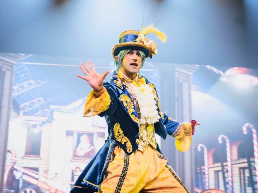 Víctor Óscar Juaranz actúa en un espectáculo circense, vestido con un colorido traje barroco azul y dorado, peluca celeste y sombrero decorado con plumas, sobre un escenario iluminado.