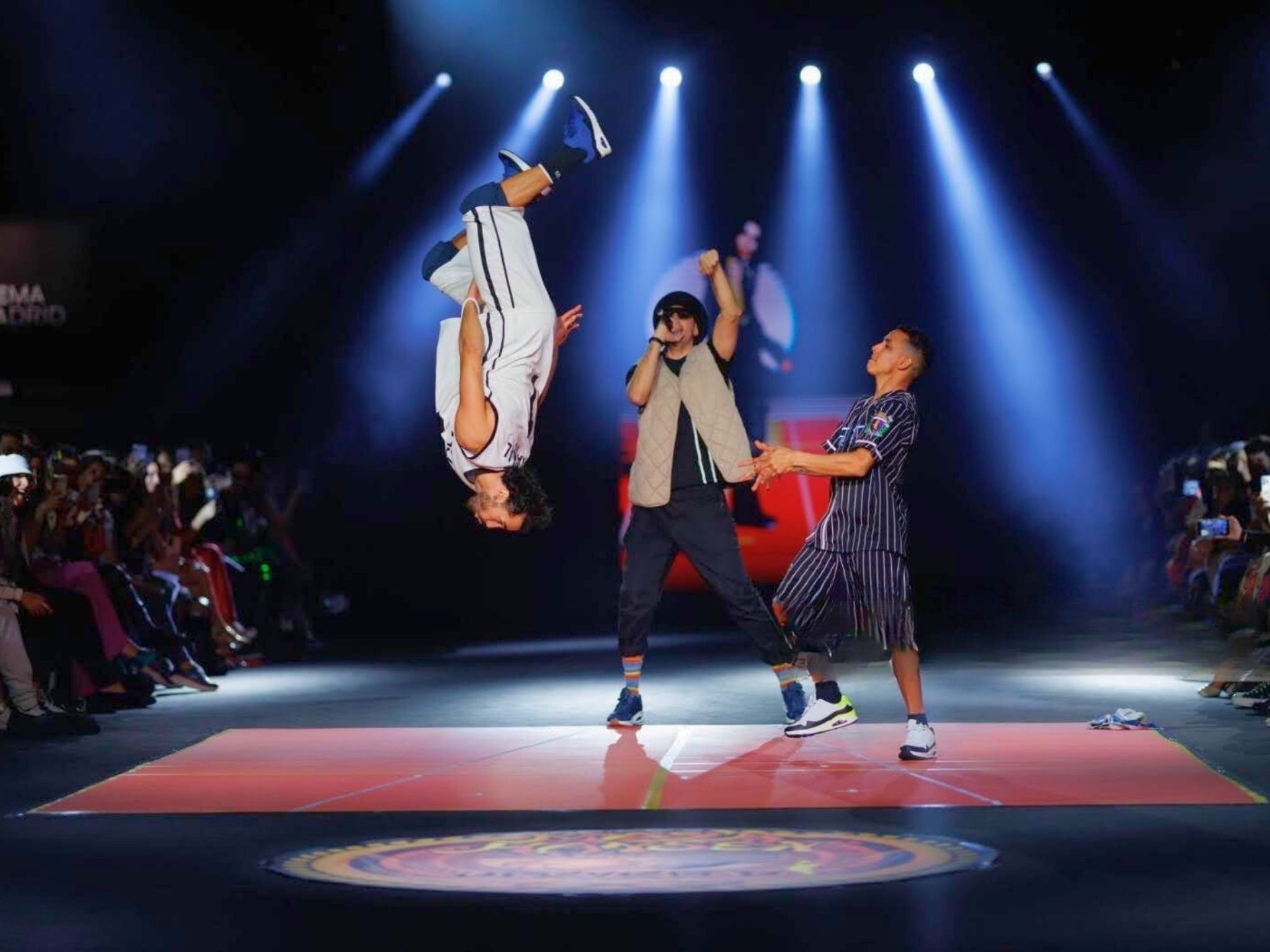 Víctor Óscar Juaranz actuando como bailarín urbano y realizando una acrobacia sobre una mano en un escenario, con otro artista rapeando y un compañero observando.