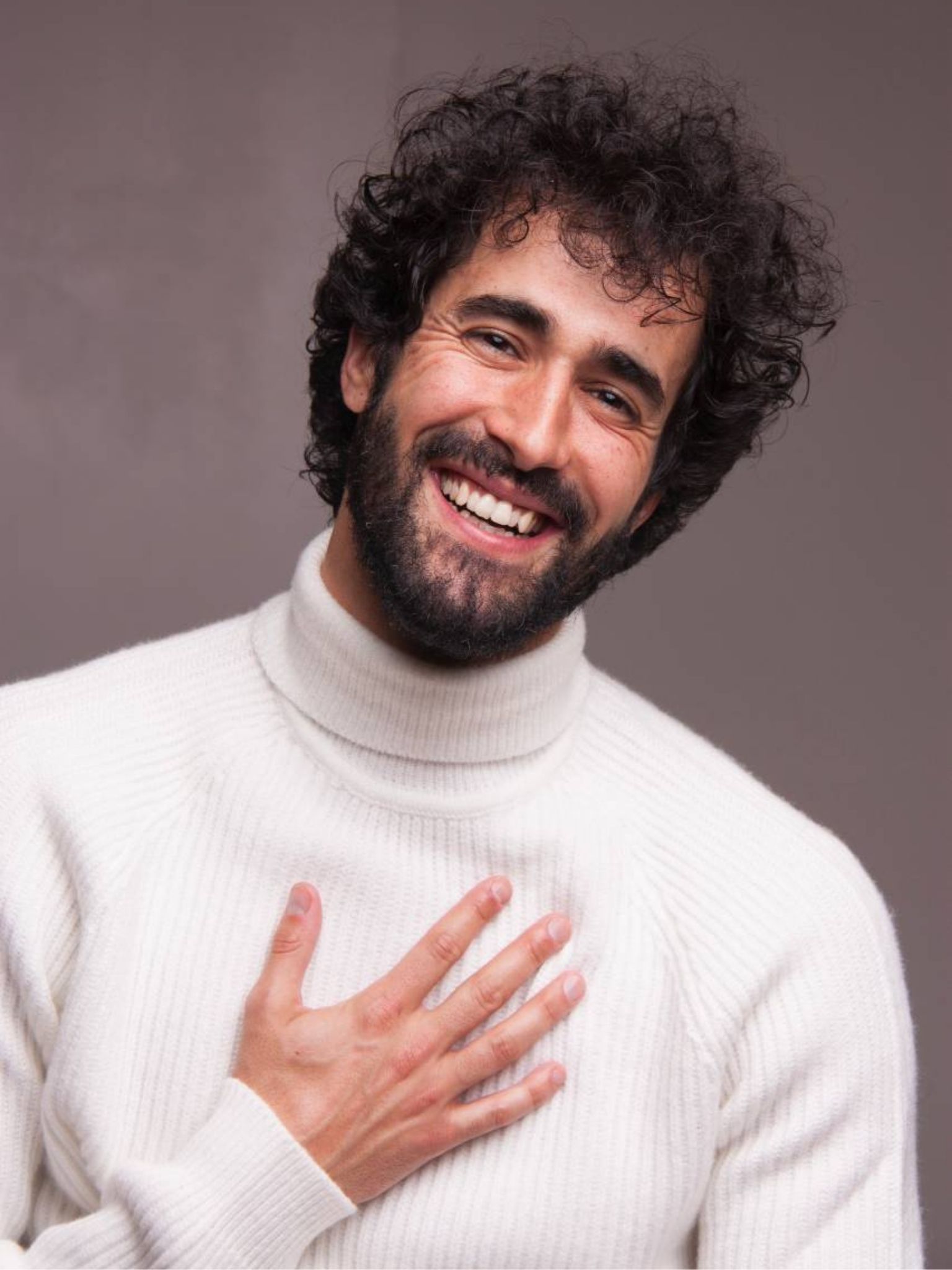 Víctor Óscar Juaranz sonríe con expresión cercana y la mano en el pecho, vistiendo un jersey blanco de cuello alto sobre fondo neutro, en un retrato cálido y expresivo.