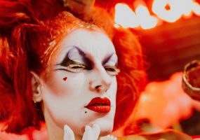 Artista caracterizada con maquillaje teatral y peluca roja, en una actuación visual.