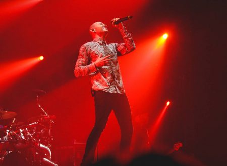 Cantante solista interpretando sobre escenario iluminado en rojo, acompañado por batería, en un concierto.