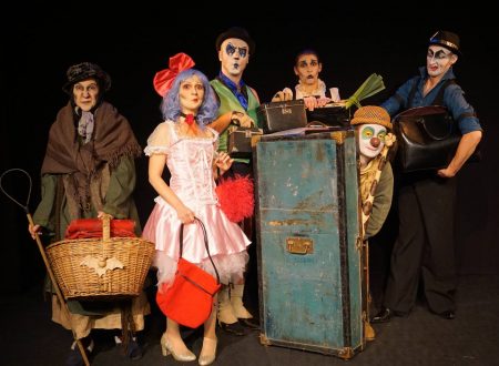 Grupo de artistas con maquillaje teatral y vestuarios coloridos posan junto a un baúl azul en escena.