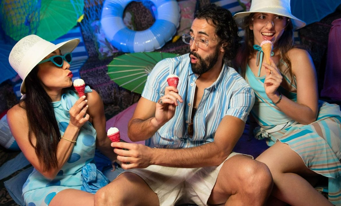 Víctor Óscar Juaranz, al centro, disfruta un brunch veraniego en Madrid junto a dos personas, todos con ropa colorida y sombreros, comiendo helados en un ambiente decorado con temática playera.