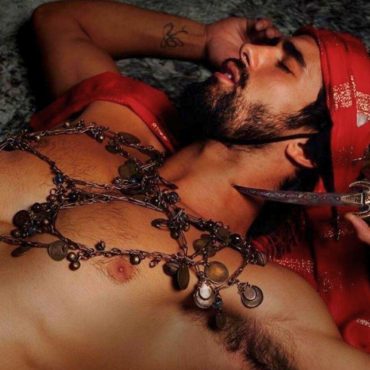 Víctor Óscar Juaranz, responsable de OjoArtísticoEvents, aparece caracterizado como Qviron, con el torso desnudo, adornado con collares rituales y una bandana roja, en una escena teatral de fuerte carga simbólica.