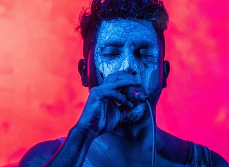 Artista masculino con el rostro pintado de blanco y azul, cantando con un micrófono frente a un fondo vibrante en tonos fucsia. Tiene los ojos cerrados y transmite intensidad emocional.