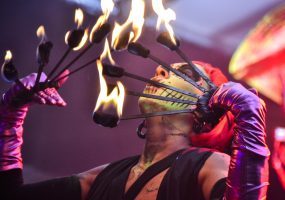 Artista con maquillaje de calavera y guantes brillantes sosteniendo abanicos de fuego encendidos durante un espectáculo escénico.
